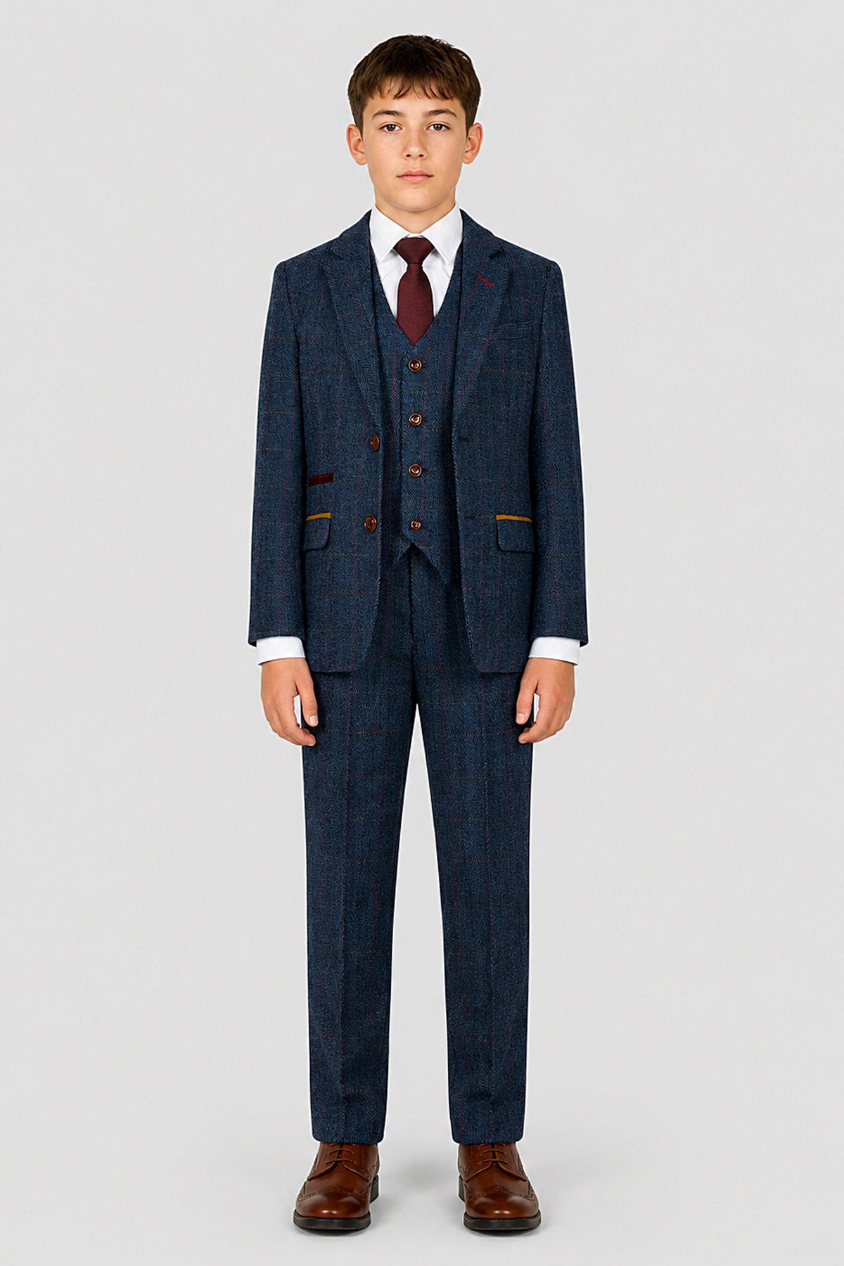 A young boy modeling a three-piece navy blue tweed suit with a subtle red and white windowpane pattern, featuring a matching vest, jacket, and trousers, styled with a white shirt, burgundy tie, and brown dress shoes against a light gray background.